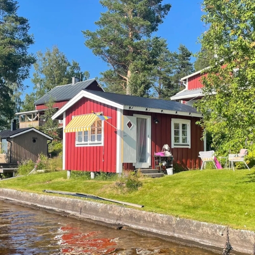 Klassisk röd sommarstuga med vit knutbräda och gul- och vitrandig fönstermarkis som ger ett snyggt solskydd.